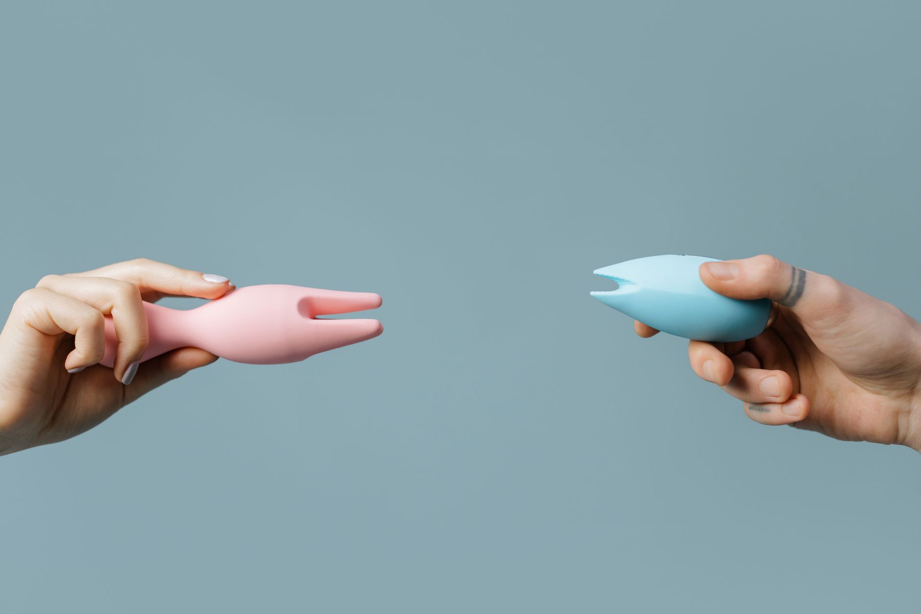 Deux mains tenant des vibrateurs en silicone rose et bleu sur fond pastel