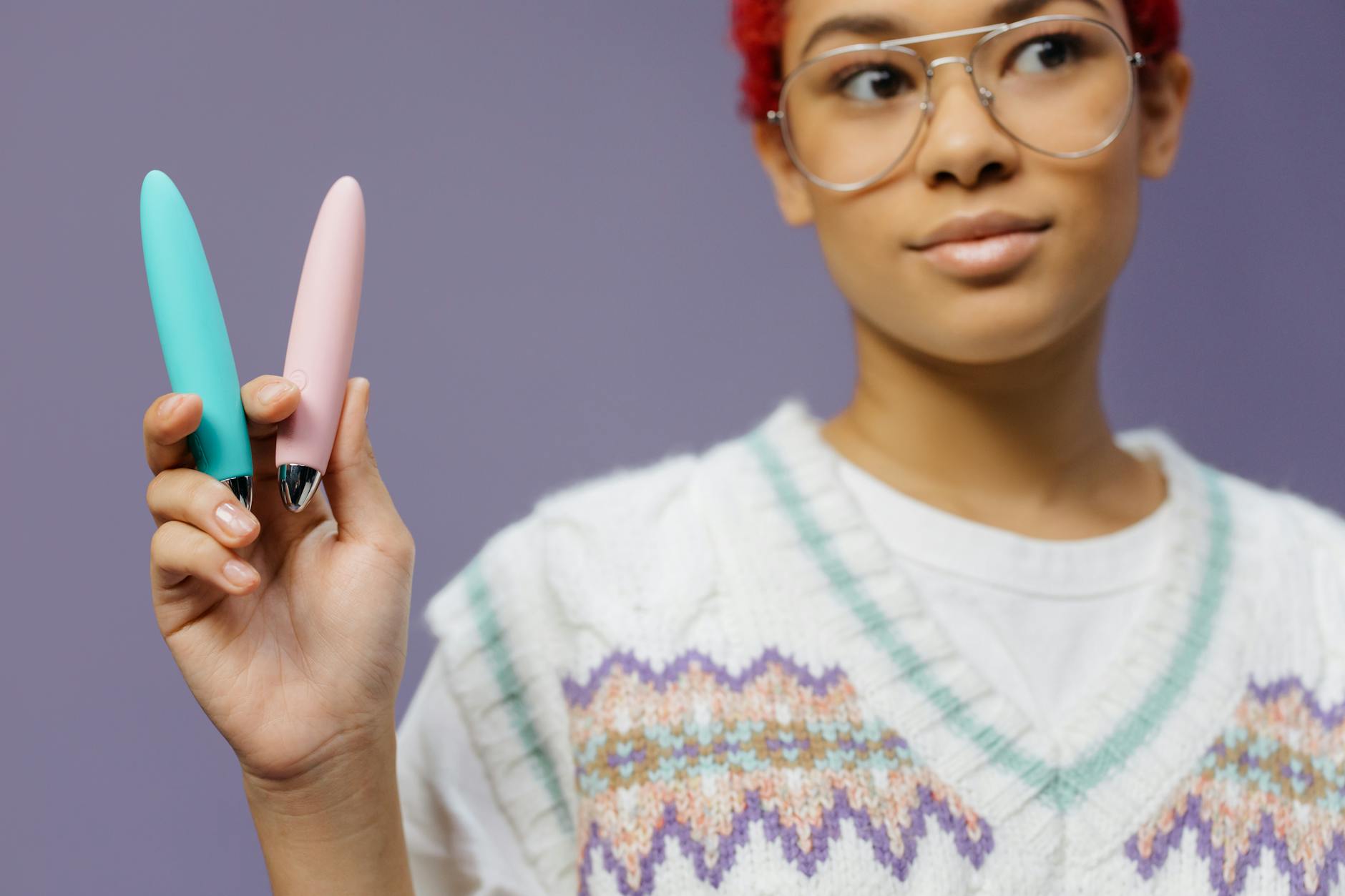 A young woman holding two colorful vibrators against a purple background, exploring pleasure at her own pace.